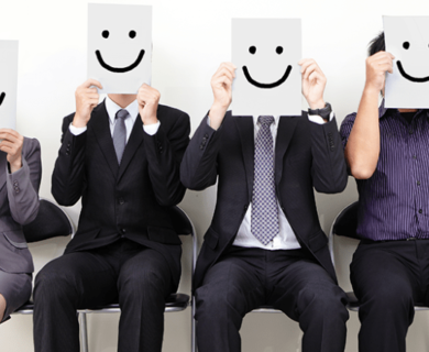 People in business attire sitting in chairs against a white wall holding cardboard smiling faces in front of their head